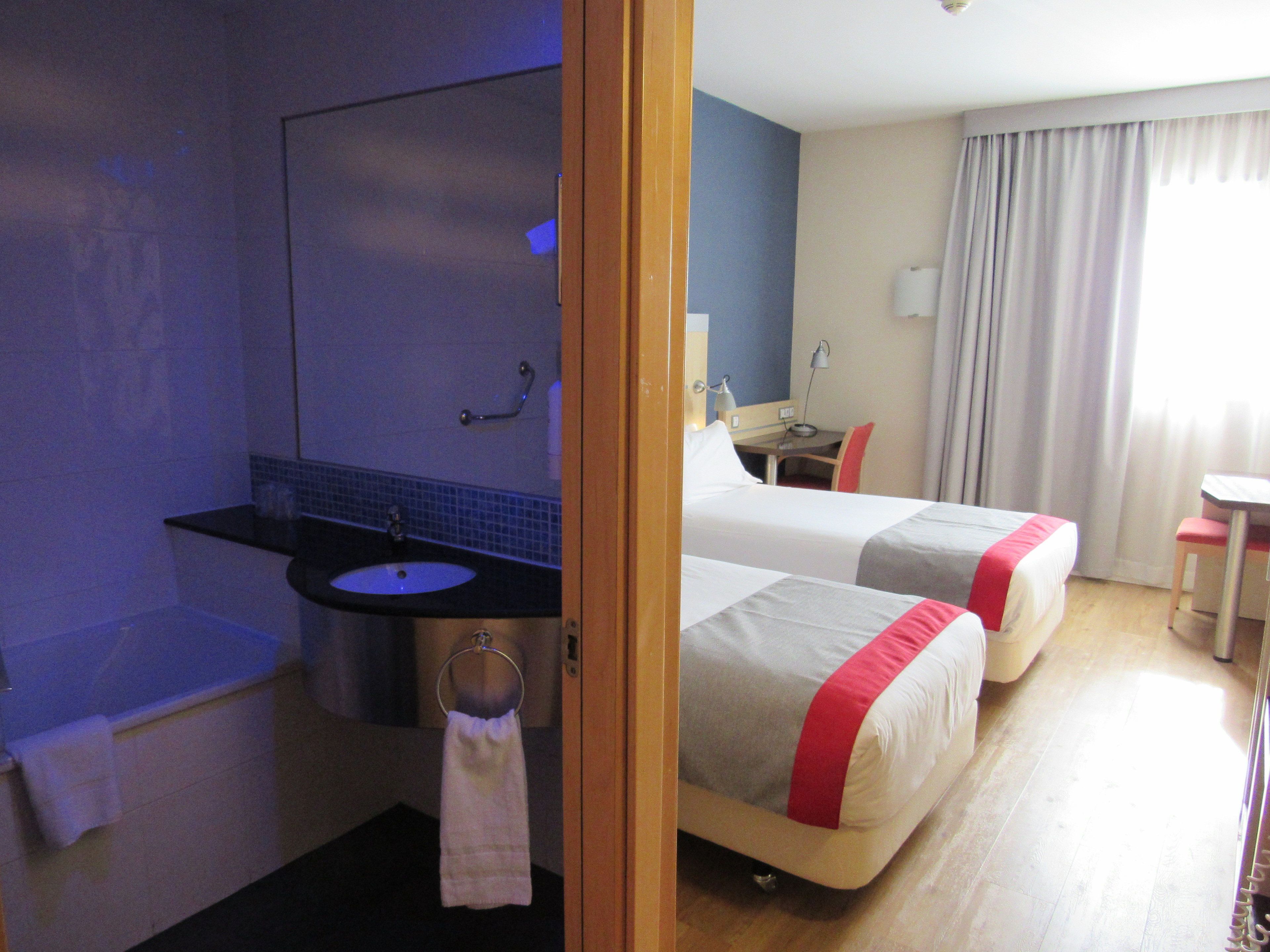 A hotel room with a bathroom, two beds, a desk, and a chair.