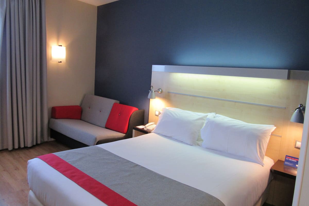Hotellihuone, jossa on suuri sänky, yöpöytä lampun kanssa, punaisilla koristetyynyillä varustettu tuoli ja ikkuna, jossa on verhot.