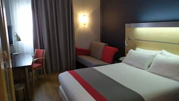Una habitación de hotel con cama, escritorio, silla y ventana con cortinas.