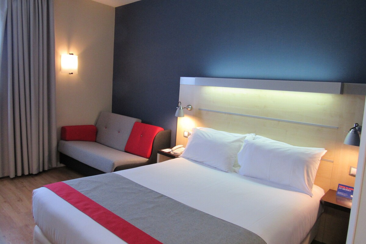 Habitación de hotel con una cama grande, una mesita de noche con lámpara, una silla con cojines rojos y una ventana con cortinas.
