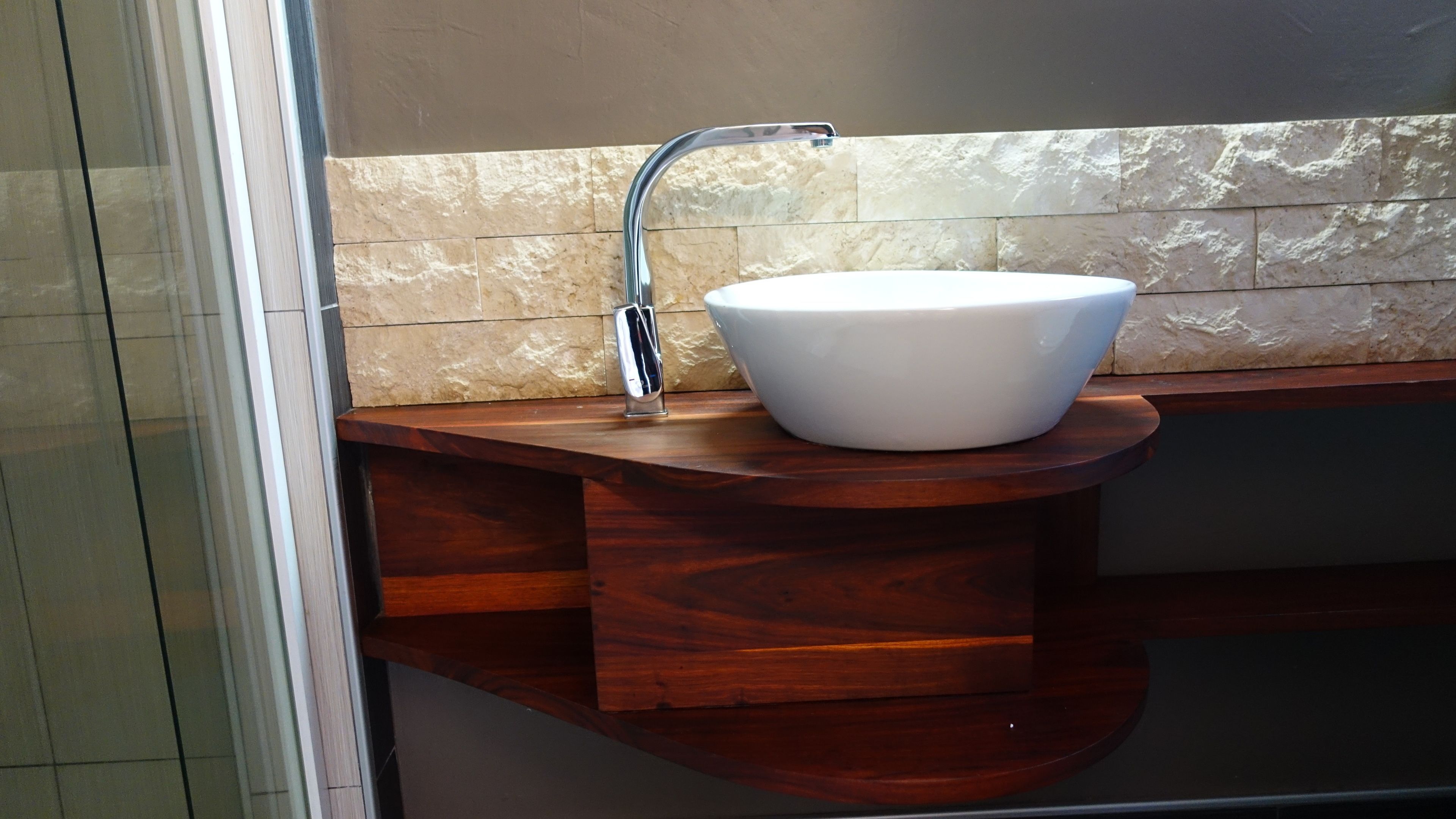 Une salle de bain avec un lavabo sur colonne blanc, un meuble-lavabo en bois et un pare-feu en pierre.