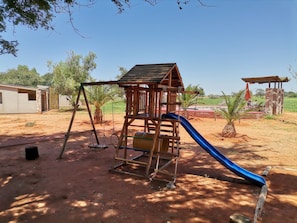 A wooden play structure with a slide, swings, and a climbing ladder in a sunny outdoor area with trees and a building in the background. - Kalahari Anib Lodge (Mariental)