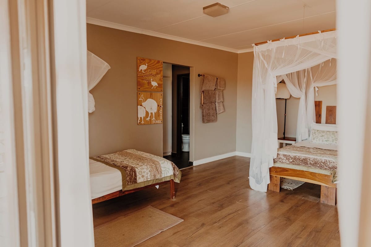 A bedroom with a wooden floor, a bed with a canopy, a smaller bed, a wall with artwork, and a doorway leading to another room.
