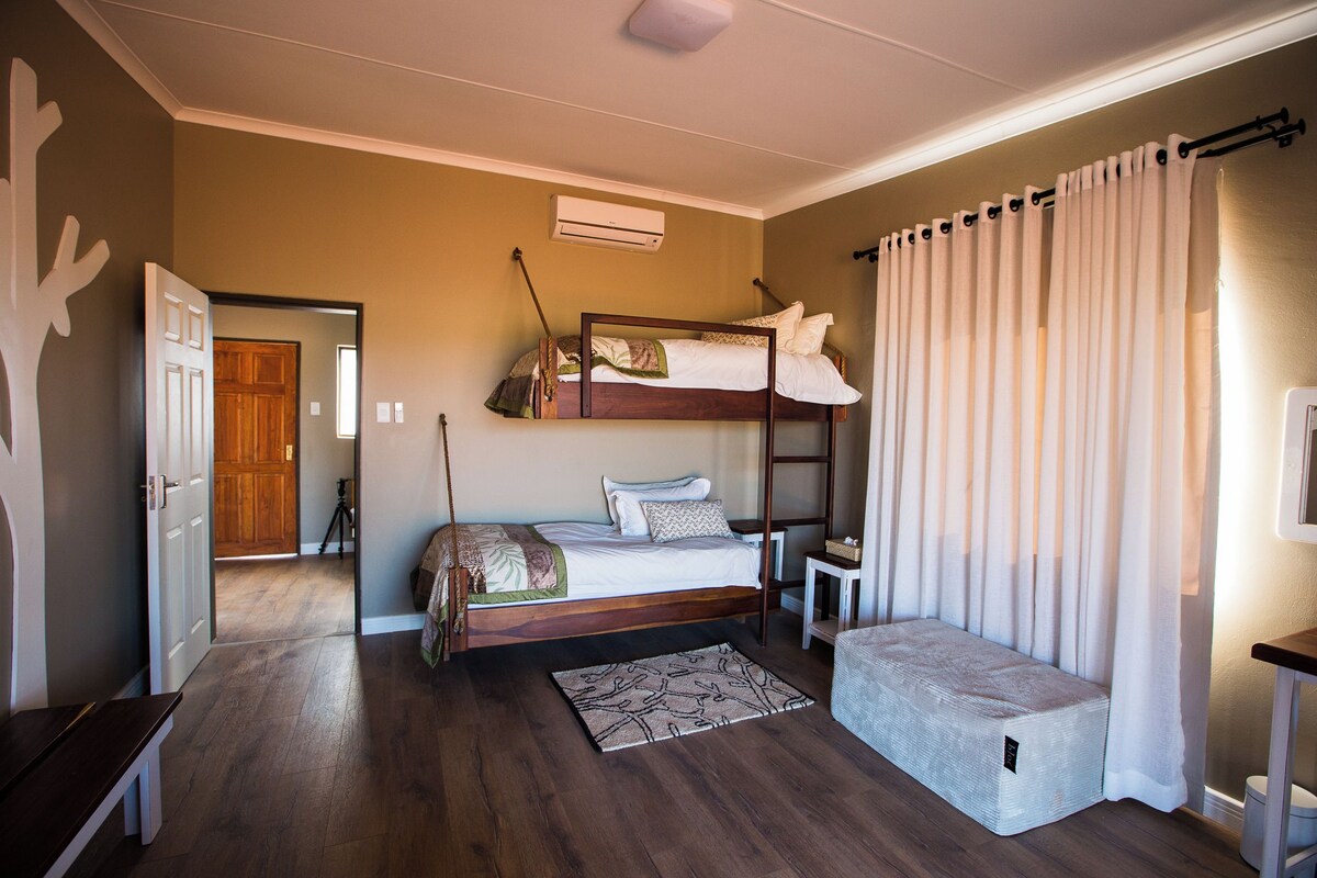 A room with a bunk bed, a bedside table, a small bench, and a window with curtains.