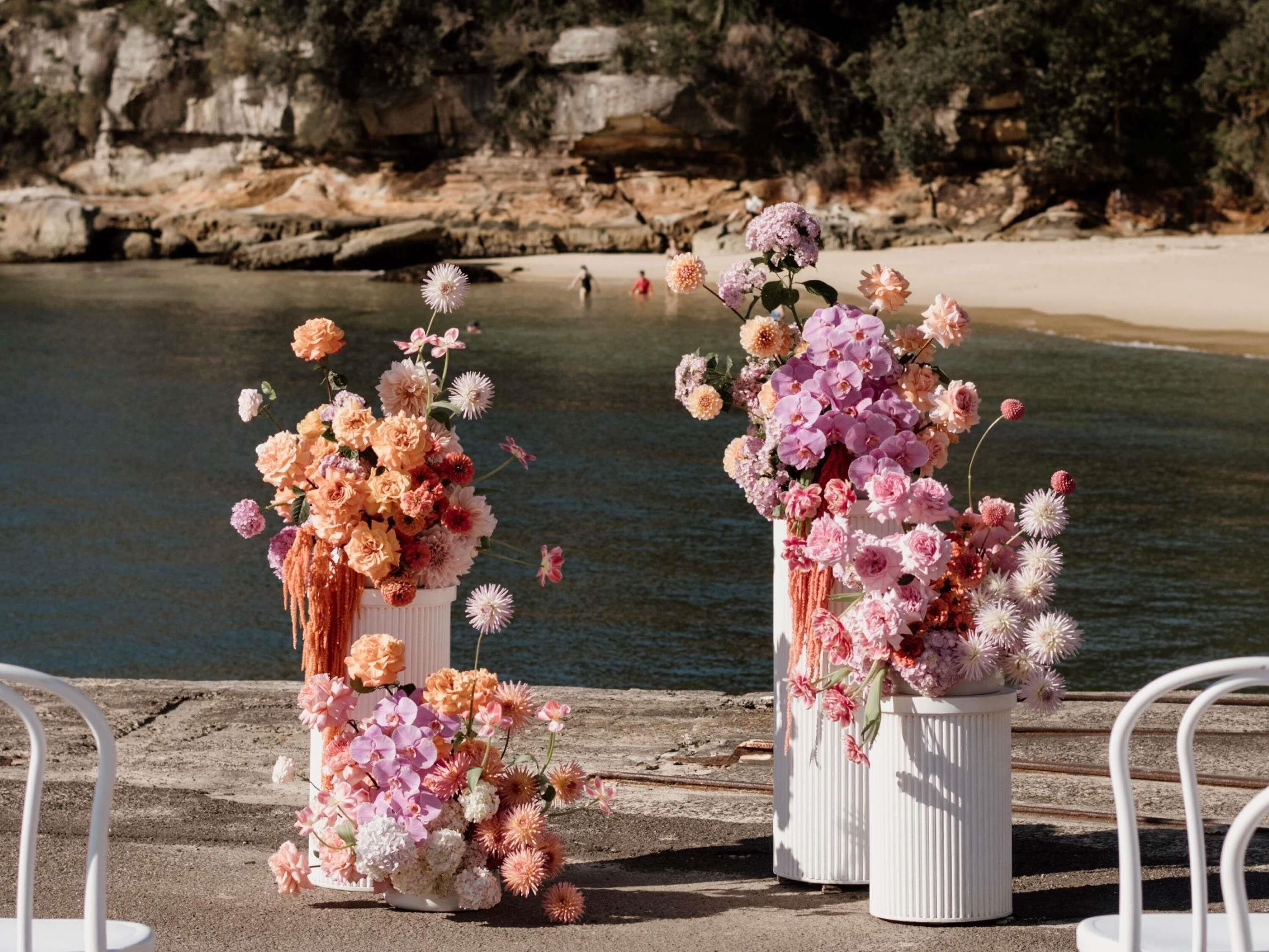 Two white flower arrangements on a concrete ledge by a body of water with rocky cliffs in the background.