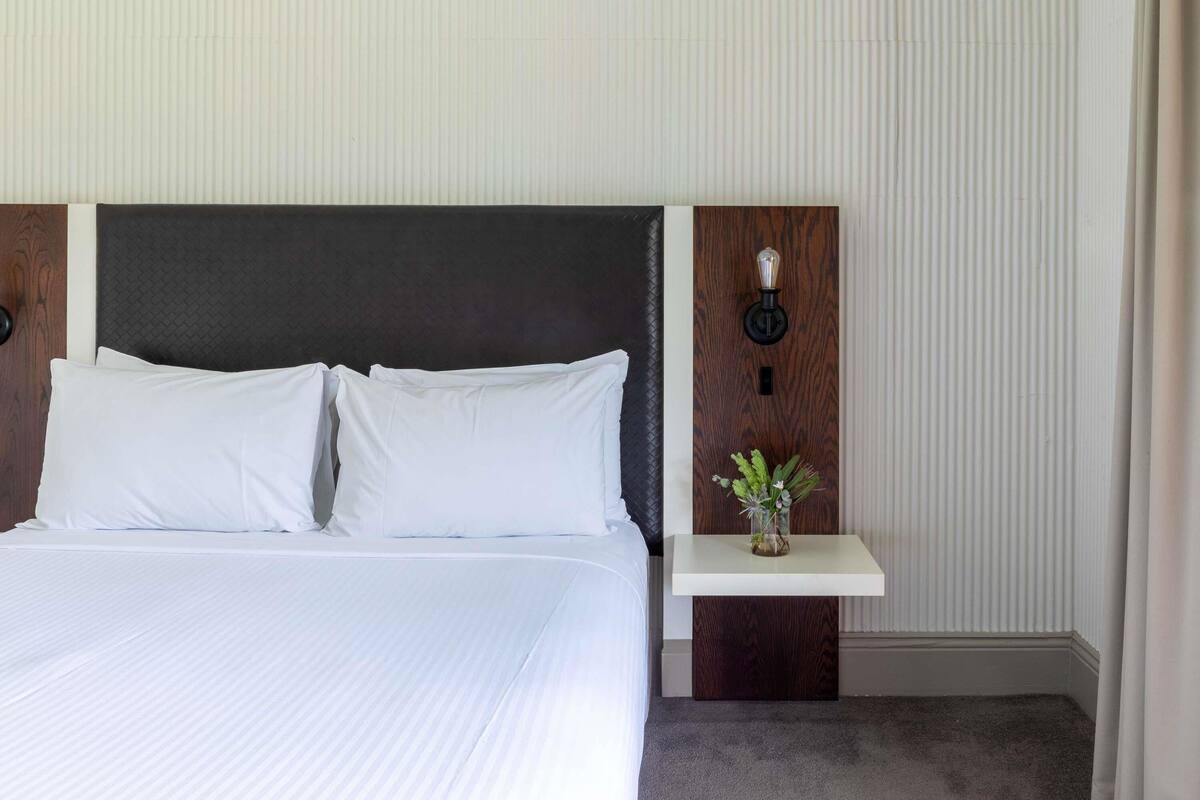 A hotel room with a large bed, a wooden headboard, a bedside table with a vase of flowers, and a wall-mounted light fixture.