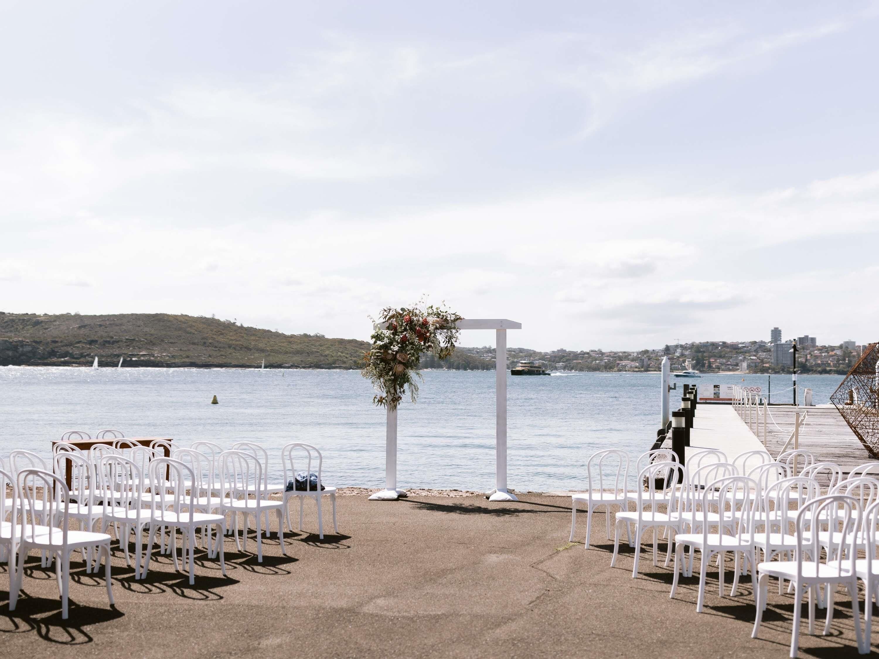 An outdoor wedding setup with white chairs arranged in rows, a white archway, and a floral decoration.
