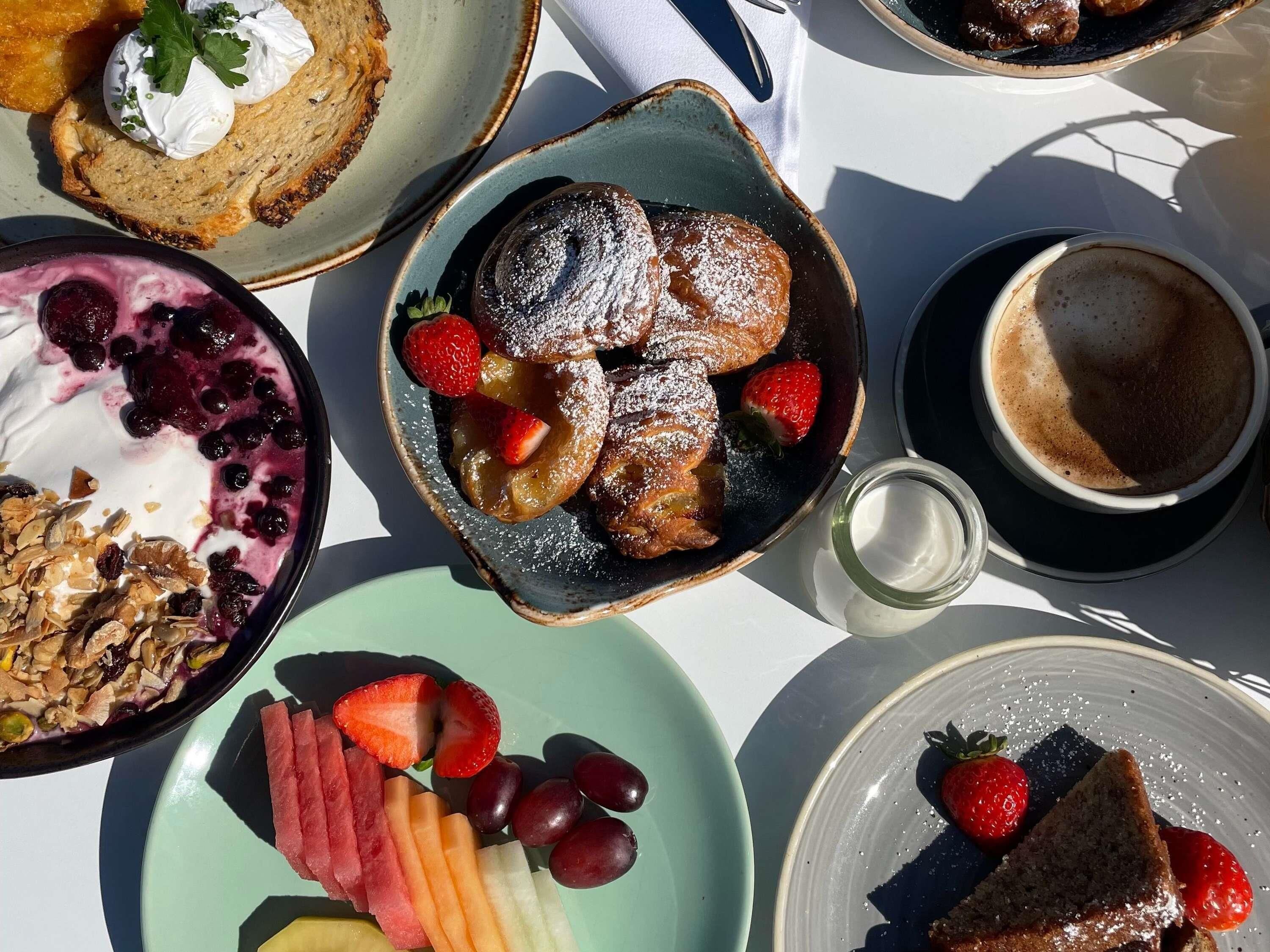 Ein Frühstückstisch mit verschiedenen Speisen, darunter Pfannkuchen, eine Schüssel Obst, eine Portion Granola und eine Tasse Kaffee.