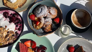 Ein Frühstückstisch mit verschiedenen Speisen, darunter Pfannkuchen, eine Schüssel Obst, eine Portion Granola und eine Tasse Kaffee.