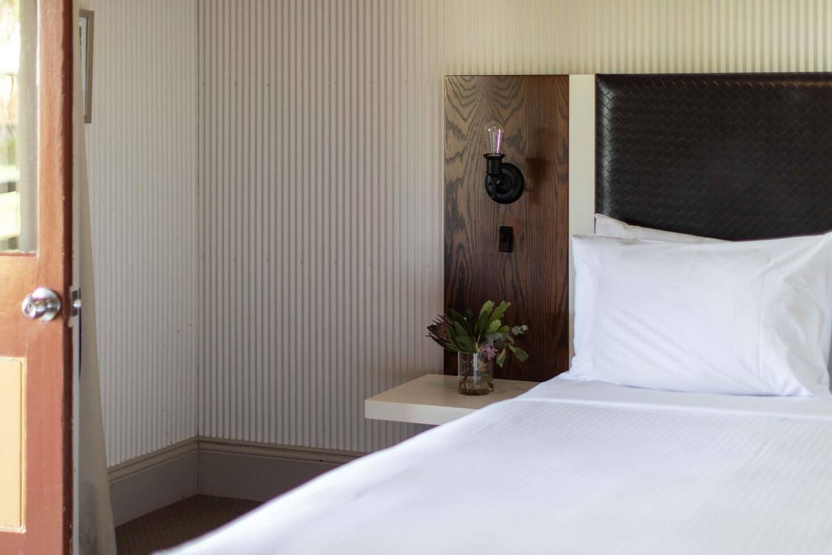 A hotel room with a bed, a nightstand with a vase of flowers, a wall-mounted light fixture, and a door.
