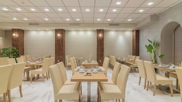 A hotel dining area with multiple tables and chairs, a patterned wall, and a ceiling with recessed lighting.