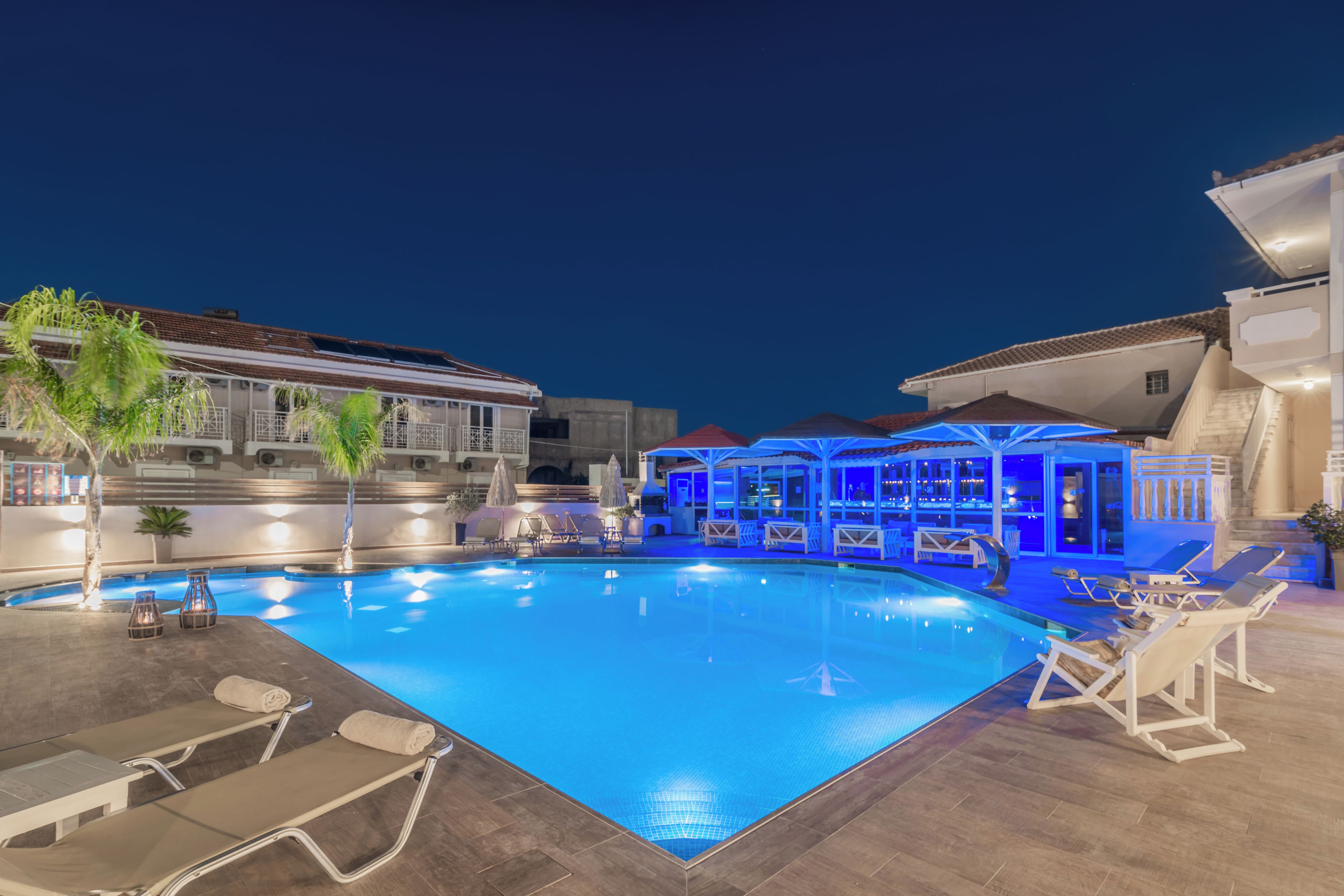 Piscina com espreguiçadeiras e guarda-sóis, cercada por edifícios com área coberta e escadaria.