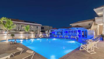 Piscina com espreguiçadeiras e guarda-sóis, cercada por edifícios com área coberta e escadaria.