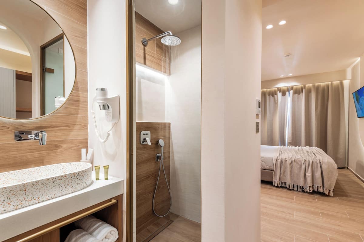 Ein modernes Badezimmer mit einem großen Spiegel, einem Waschbecken mit Pflegeprodukten, einer Dusche mit Duschbrause und einem Bett mit Decke.