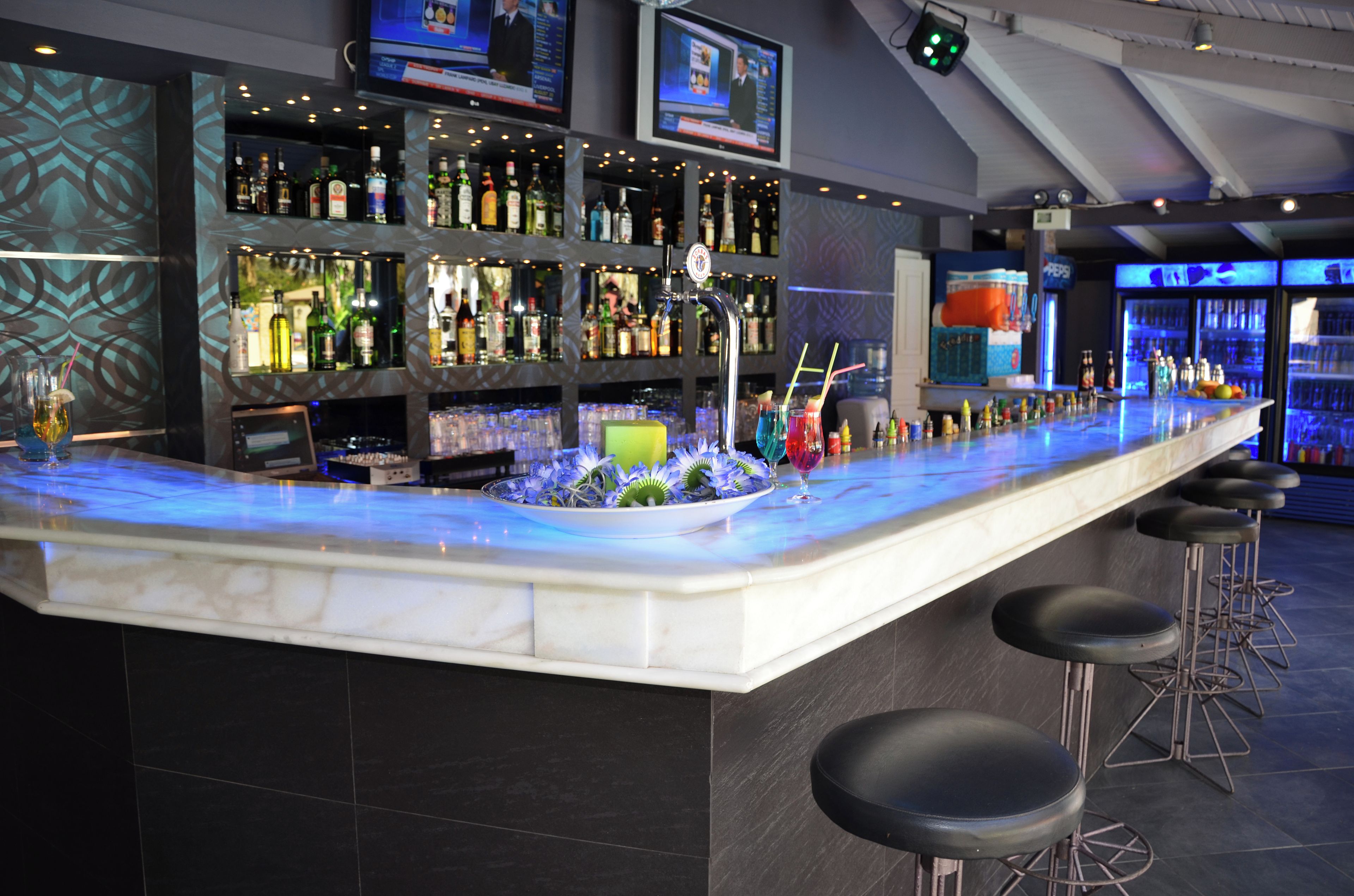 Zona de bar con mostrador de mármol, varias estanterías llenas de botellas y taburetes.