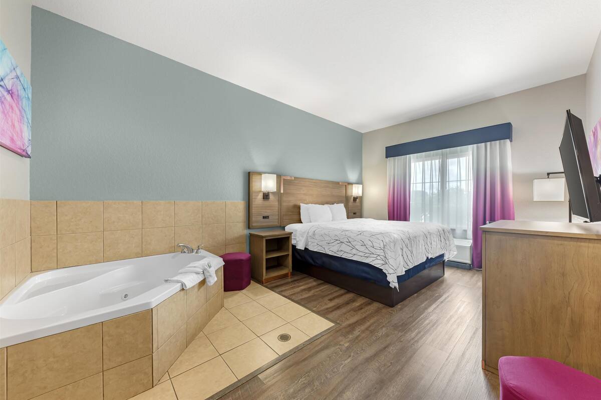 Habitación de hotel con una bañera grande, una cama de madera con cabecera azul, un escritorio de madera y una ventana con cortinas moradas.