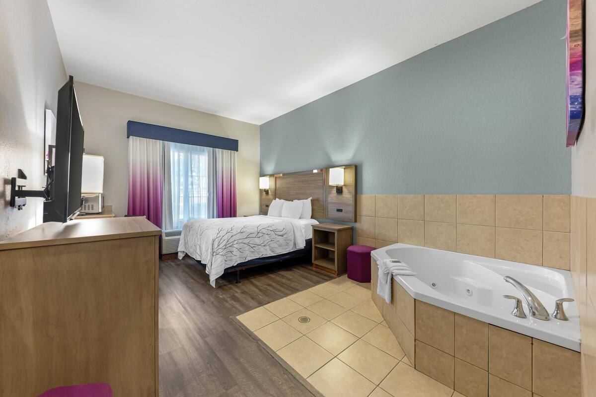 Habitación de hotel con cama, mesita de noche, lámpara y baño con bañera y lavamanos.