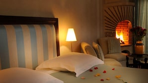 Quarto de hotel com cama, almofadas, abajur, lareira de tijolos e um vaso com flores.