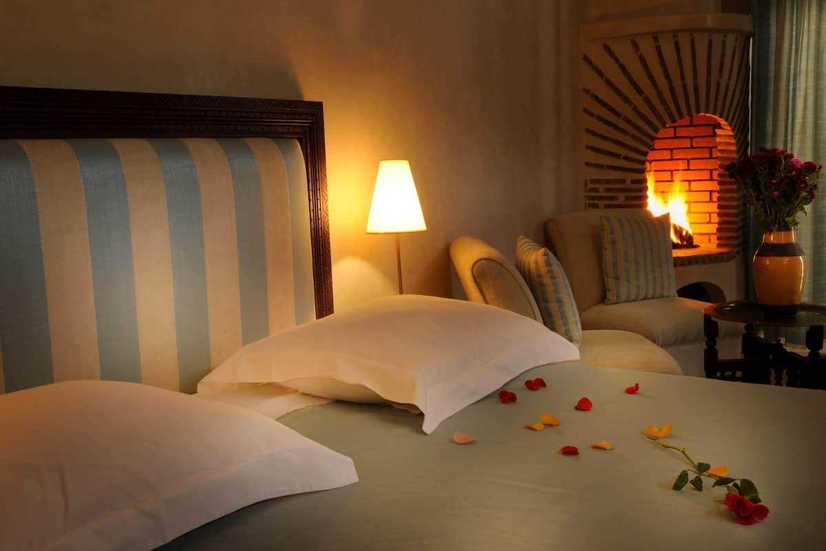 A hotel room with a bed, pillows, a lamp, a brick fireplace, and a vase with flowers.