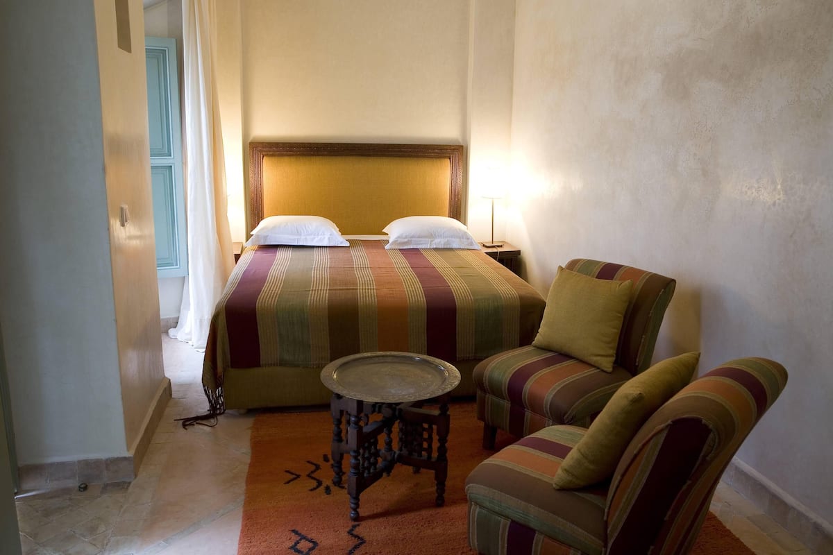 A hotel room with a bed, a chair, a small table, and a lamp.