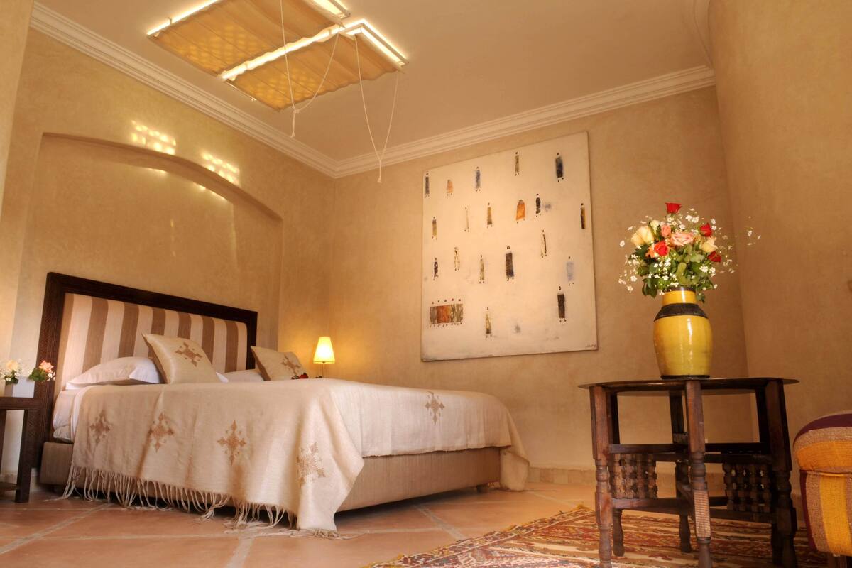 A hotel room with a large bed, a nightstand, a lamp, a vase with flowers, and a wall with artwork.