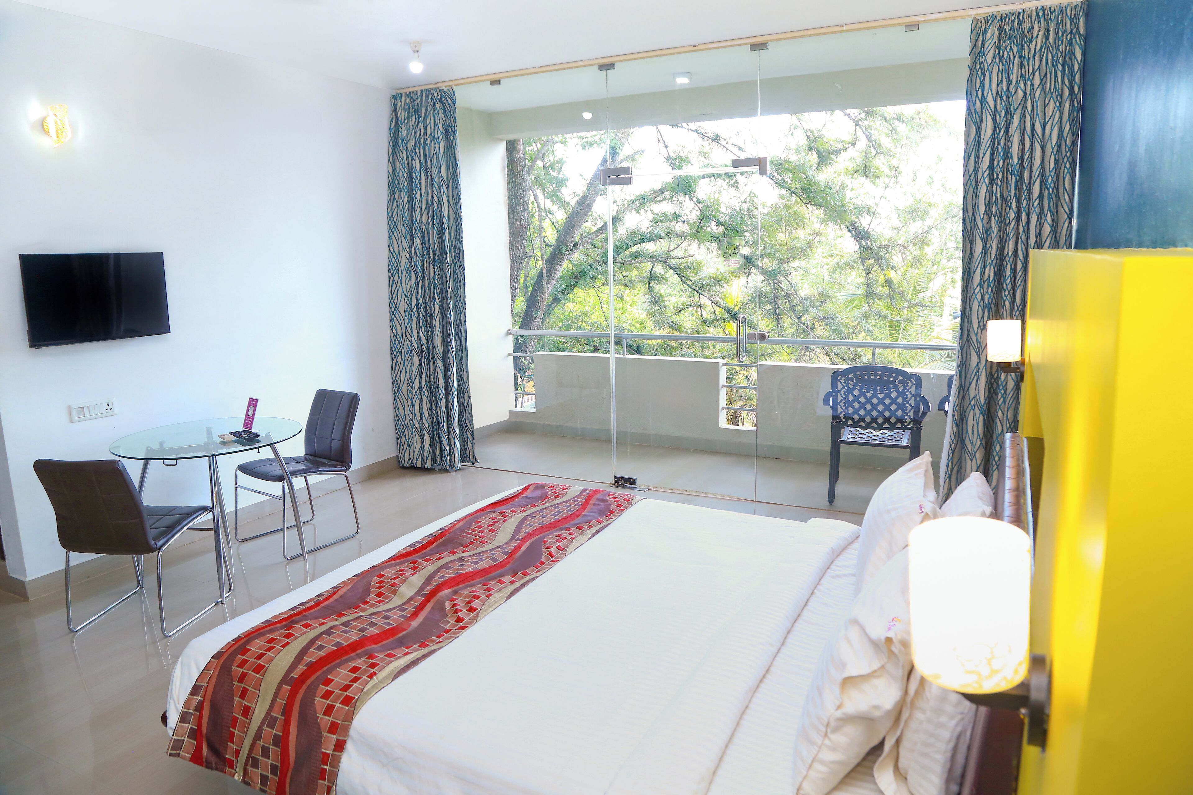 Hotelkamer met een bed, een bureau met stoel, een aan de muur bevestigde televisie, en een balkon met uitzicht op bomen.