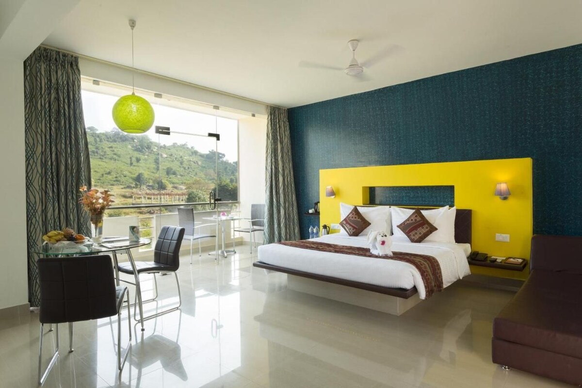 A modern hotel room with a large bed, a balcony with a view of the hills, a dining area with a table and chairs, and a bright yellow accent wall.