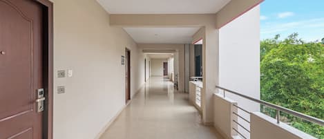 A long, narrow corridor with a wooden door on the left, a white wall with a door number, and a balcony with a metal railing on the right.