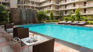 A pool area with lounge chairs and tables, a waterfall feature, and a multi-story building with balconies. - Sterling Yelagiri (Tirupattur)