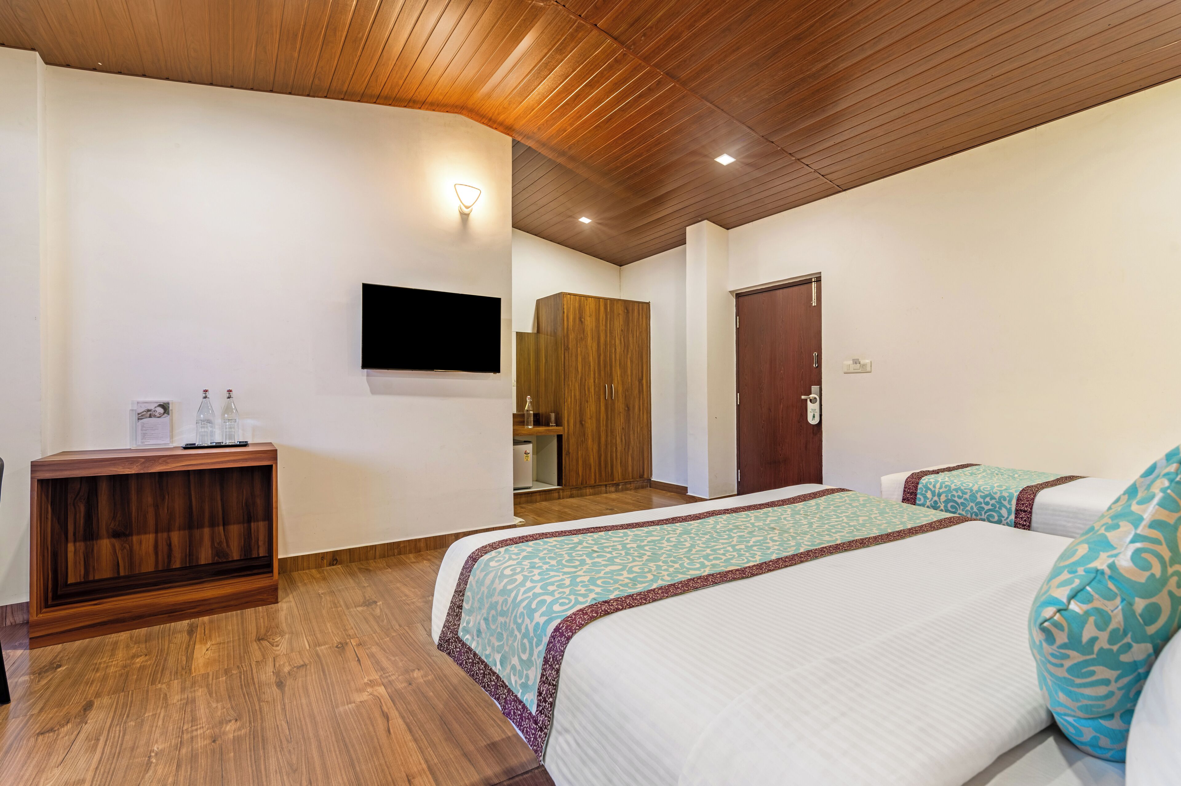 Ein Hotelzimmer mit zwei Betten, einem Holzkonsolentisch, einem an der Wand montierten Fernseher und einer Holzbalkendecke.
