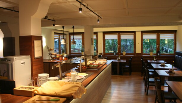 A restaurant with a long counter, wooden tables, and chairs. There are potted plants and a refrigerator on the left side. - Gästehaus Elisabeth (Tulfes)
