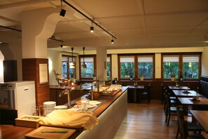 A restaurant with a long counter, wooden tables, and chairs. There are potted plants and a refrigerator on the left side. - Gästehaus Elisabeth (Tulfes)