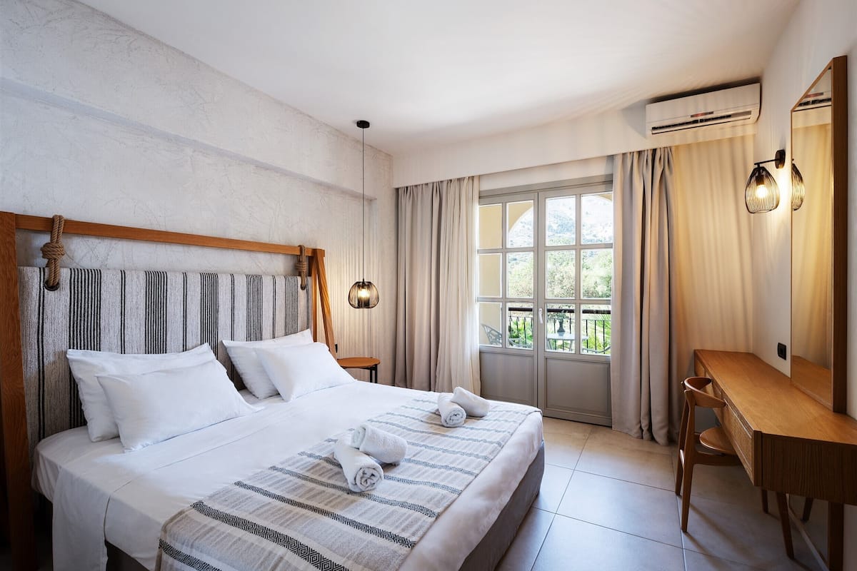A hotel room with a large bed, a wooden desk, and a view of the outdoors through a large window.