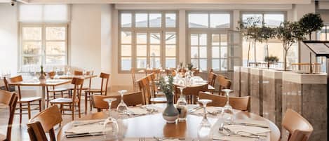 Un restaurante con mesas redondas, sillas de madera y ventanas amplias.