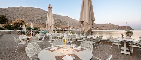 Un restaurante frente al mar con terraza, mesas y sombrillas, con vistas a un mar tranquilo y colinas rocosas.