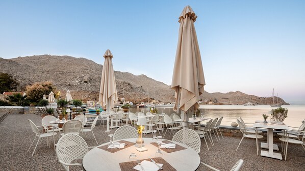 A seaside restaurant with outdoor seating, tables, and umbrellas, overlooking a calm sea and rocky hills. - Pedi Beach Hotel (Symi)