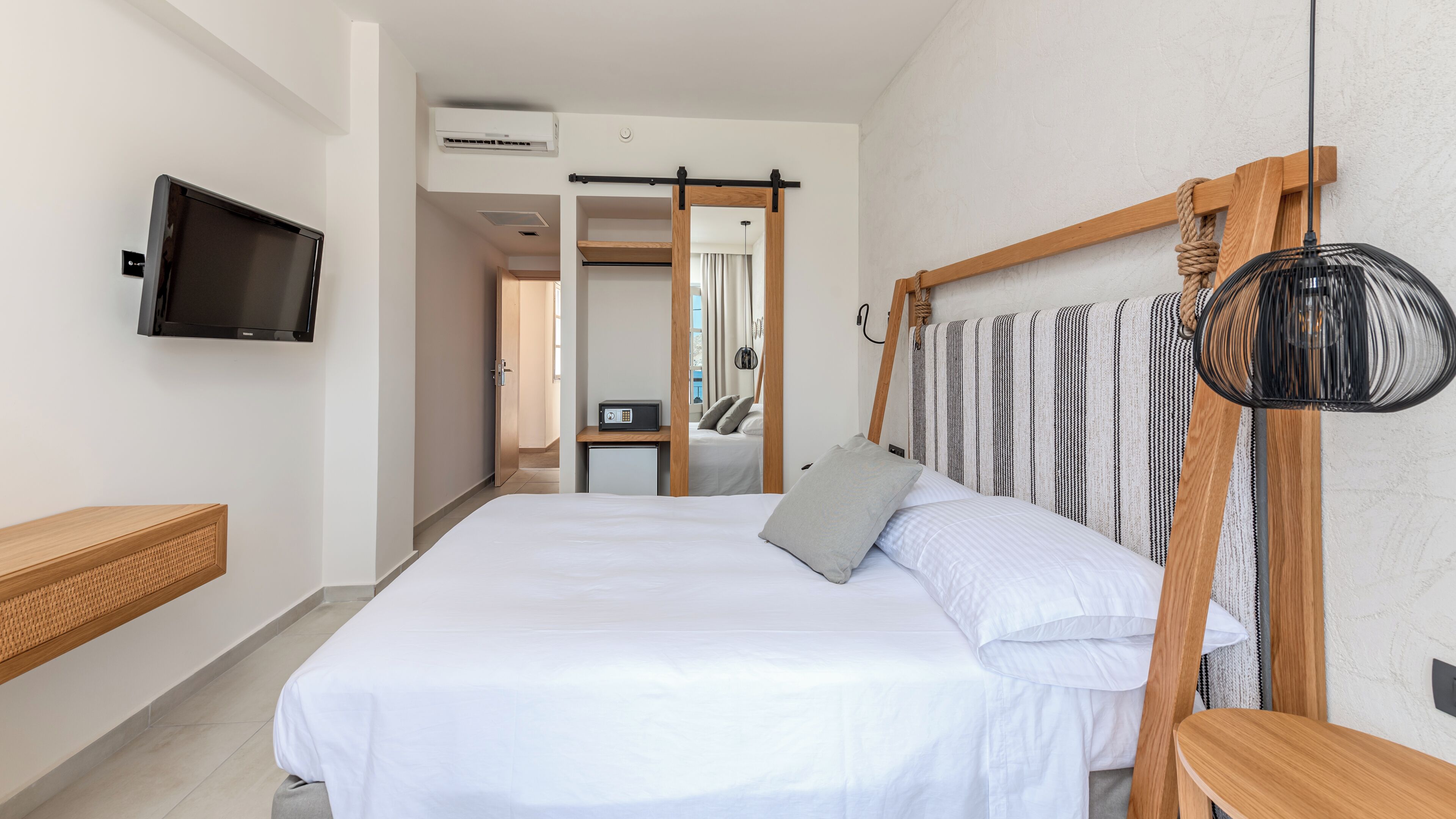 Quarto de hotel com cama, televisão presa na parede, mesa de cabeceira de madeira e um espelho grande.