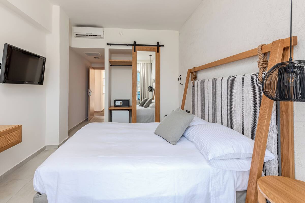A hotel room with a bed, a TV mounted on the wall, a wooden bedside table, and a large mirror.