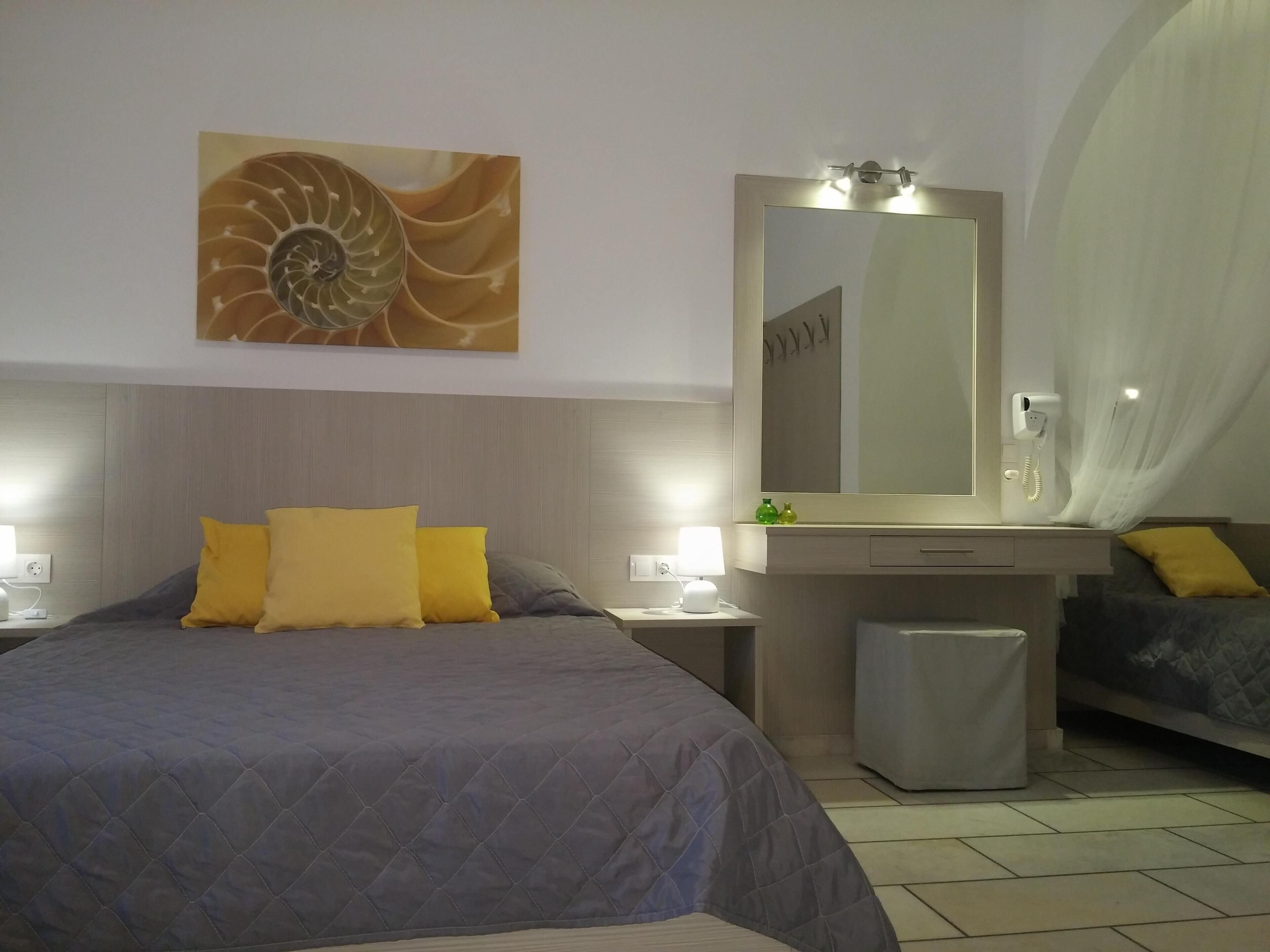 Habitación de hotel con una cama grande, una mesita de noche con lámpara, un tocador con espejo y un pequeño cubo de basura.