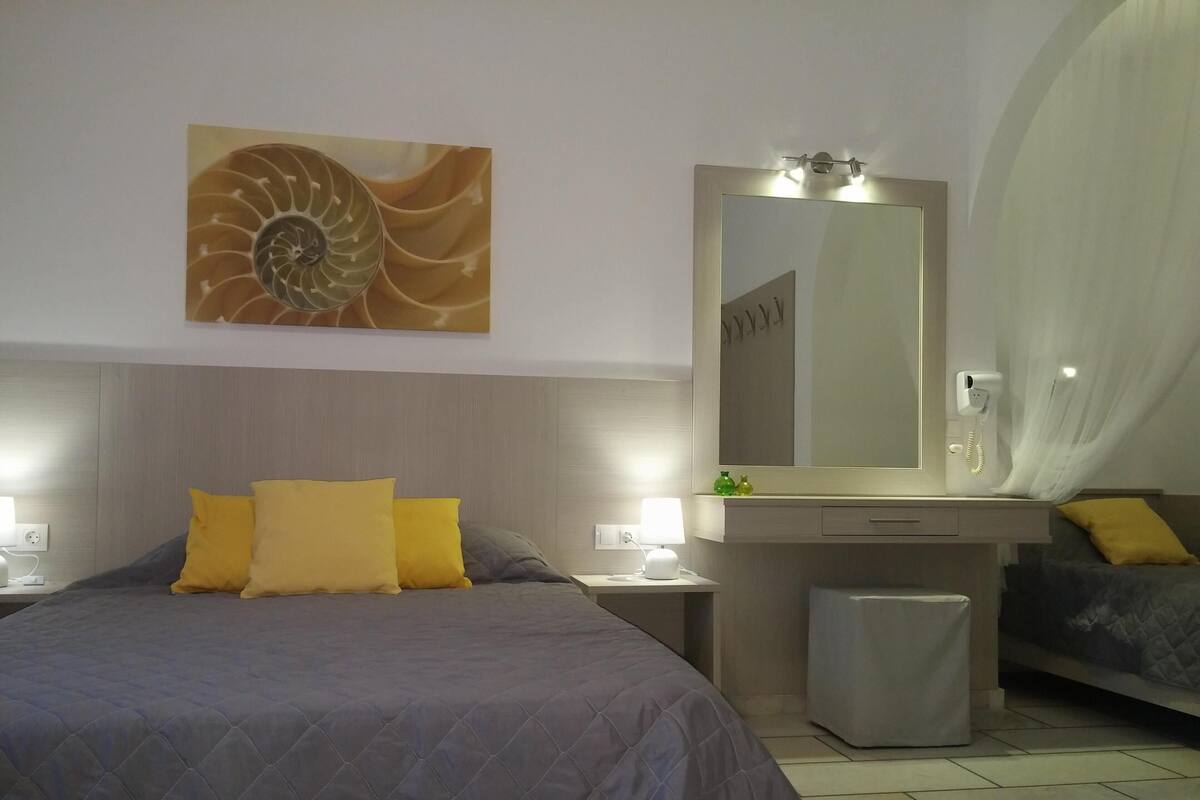 Habitación de hotel con una cama grande, una mesita de noche con lámpara, un tocador con espejo y un pequeño cubo de basura.