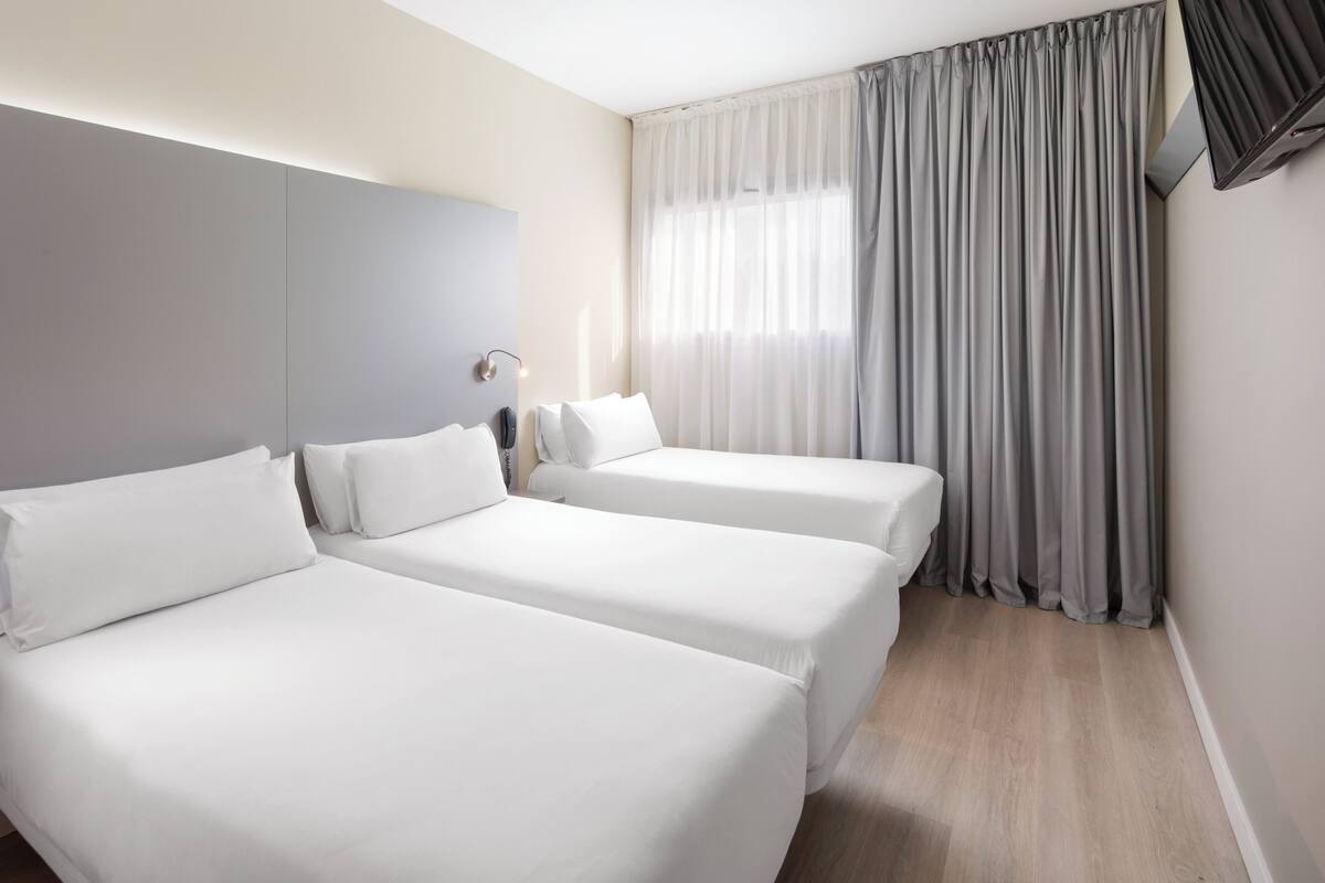 Une chambre d’hôtel avec deux lits simples, une table de chevet avec une lampe, une grande fenêtre avec des rideaux et un téléviseur à écran plat fixé au mur.