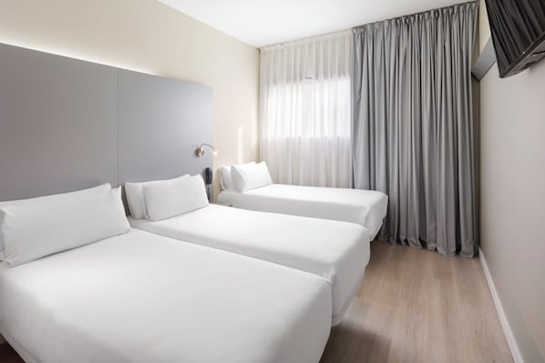 A hotel room with two single beds, a bedside table with a lamp, a large window with curtains, and a flat-screen TV mounted on the wall.