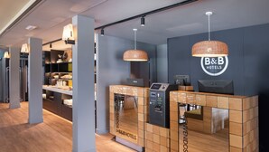 Un espace d’accueil au design moderne, comprenant un comptoir d’accueil, un kiosque numérique et une enseigne pour la chaîne B&B Hotels.