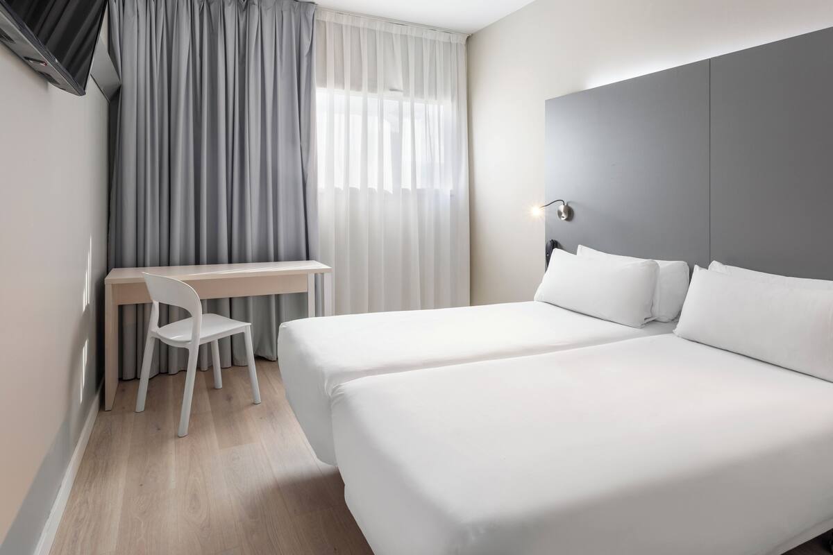 Une chambre d’hôtel avec deux lits, un bureau, une chaise et une grande fenêtre avec des rideaux.