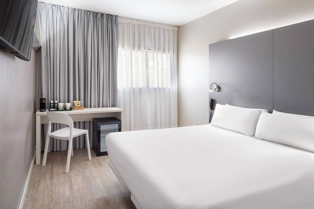 Une chambre d’hôtel avec un lit, un bureau et une chaise, une télévision et une fenêtre avec des rideaux.