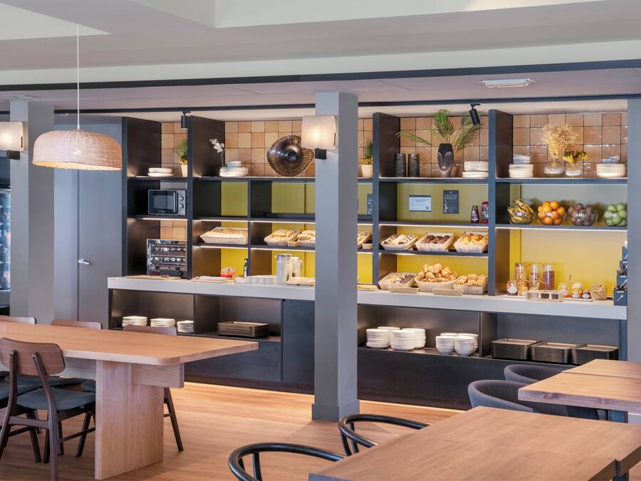 Un café moderno con mesas y sillas de madera, un mostrador de buffet con distintos alimentos y un refrigerador lleno de bebidas.