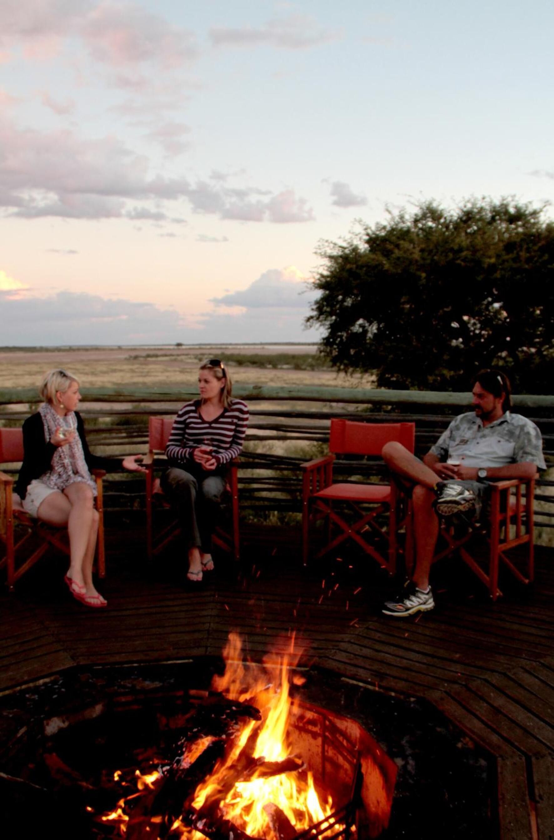 Three people seated around a campfire on a wooden deck with a view of the landscape.