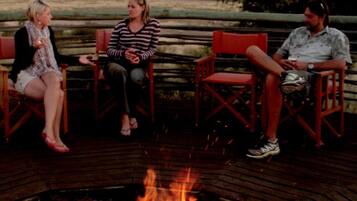 Three people seated around a campfire on a wooden deck with a view of the landscape.