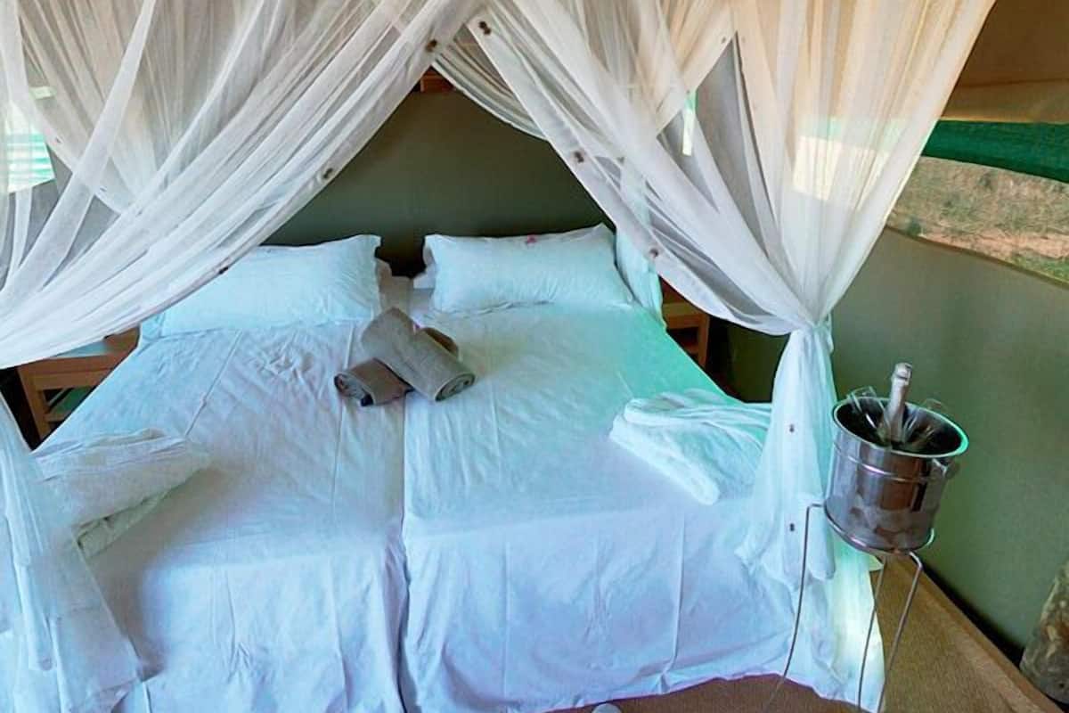 Una cama con sábanas blancas y un mosquitero. Hay un balde de metal sobre un soporte al lado de la cama.