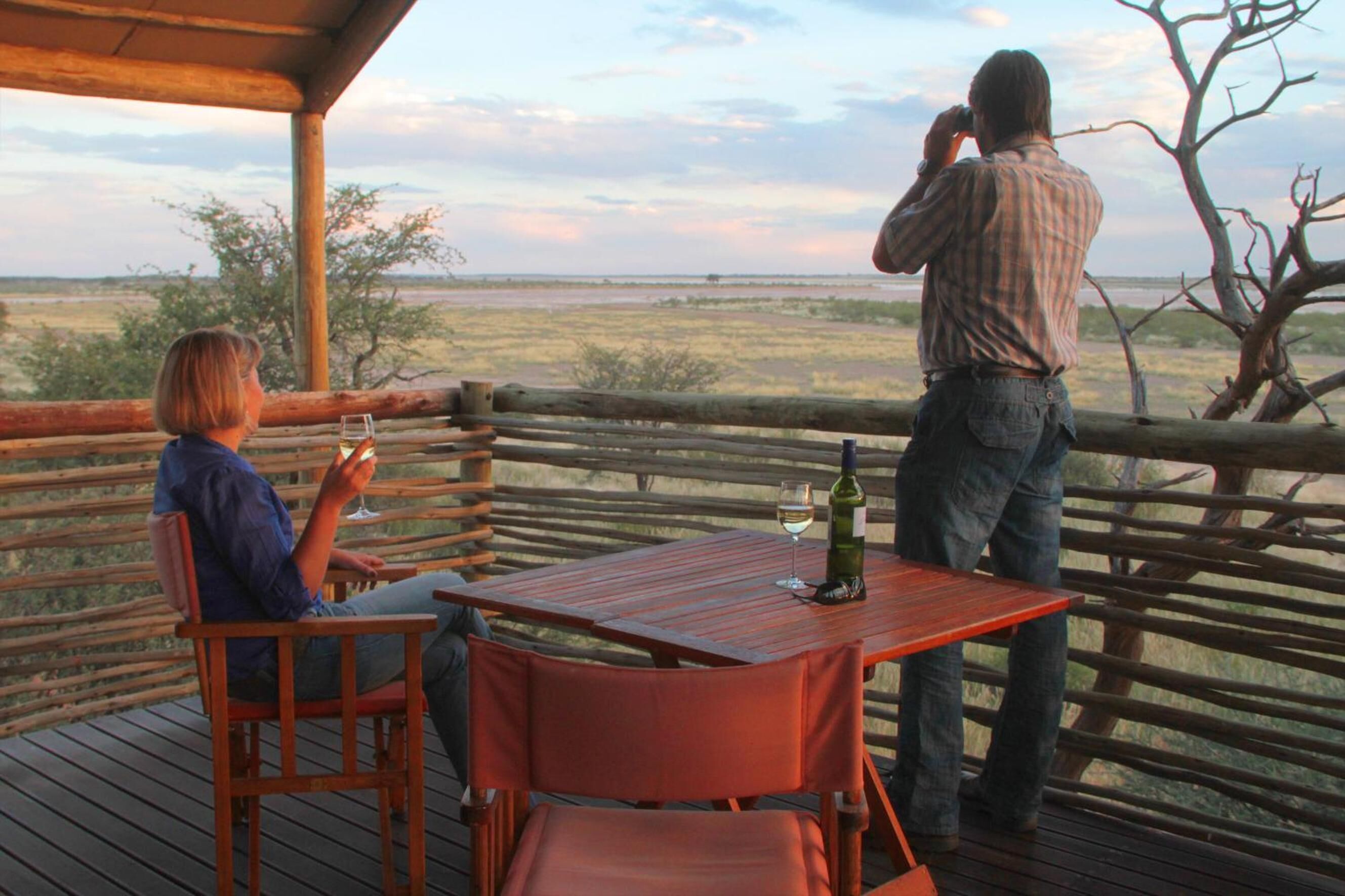 A wooden deck with two chairs and a table, a bottle of wine, and a glass on the table.